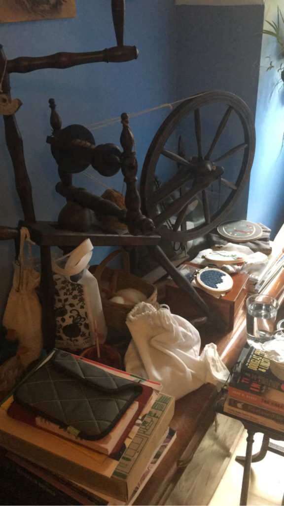 Jumble of drop spindles and fiber, books, and embroidery projects around a spinning wheel. Everything is sitting on a wooden blanket chest that is barely visible. Bright light is coming in from a window just beyond the right edge of the image.