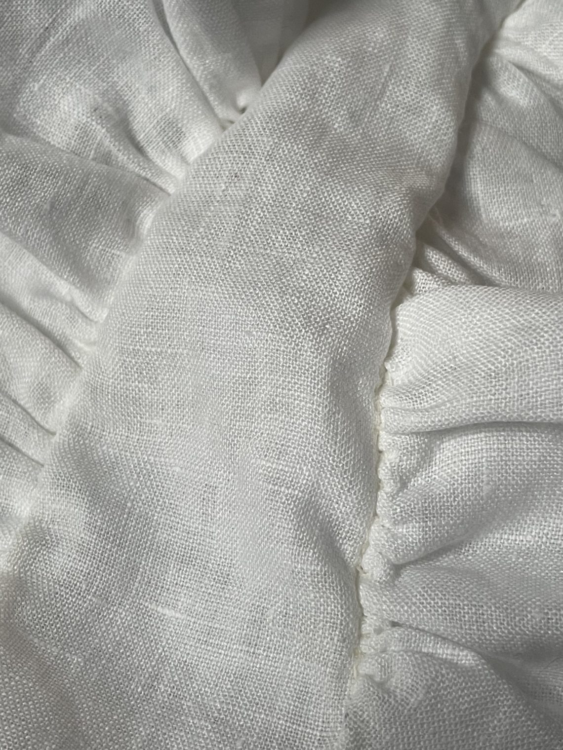 Closeup of white linen 18th century cap. Smooth headpiece is centered in the image with gathered areas visible to both sides.