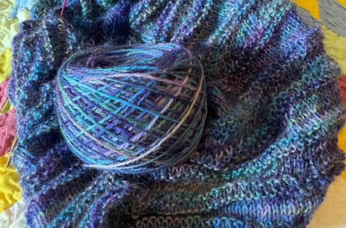 blue and purple ball of yarn sitting on top of an unfinished hand knit shawl. Knitting is sitting on a brightly colored quilt.