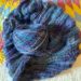 blue and purple ball of yarn sitting on top of an unfinished hand knit shawl. Knitting is sitting on a brightly colored quilt.
